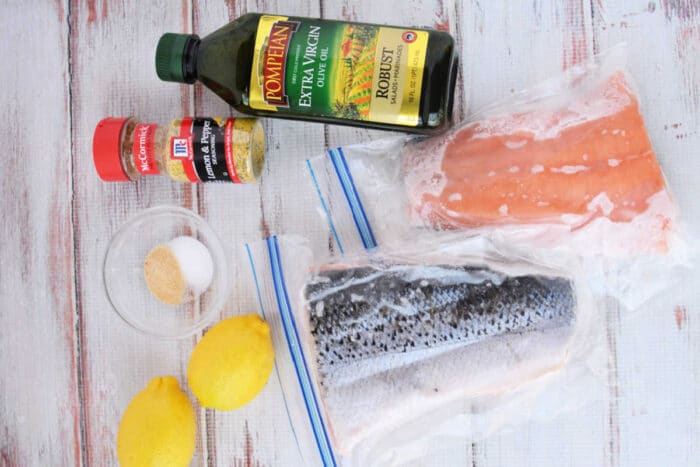 Two raw or frozen salmon fillets, olive oil, spices, lemons, and seasonings are arranged on a white wood surface, ready to be cooked in the oven.