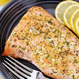 Baked frozen salmon fillet on a black plate with lemon slices and a fork, showing how deliciously simple it is to prepare frozen salmon in the oven.