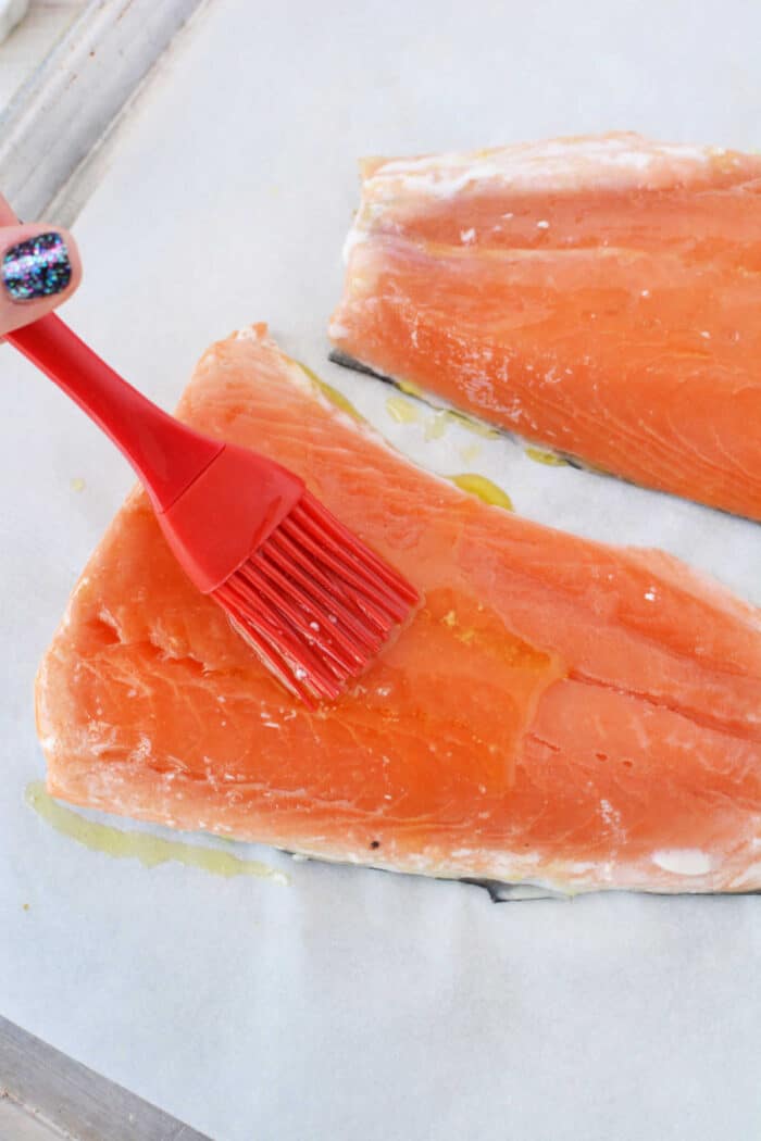 A hand brushes oil onto frozen salmon fillets with a red silicone brush on parchment paper, preparing them for the oven.