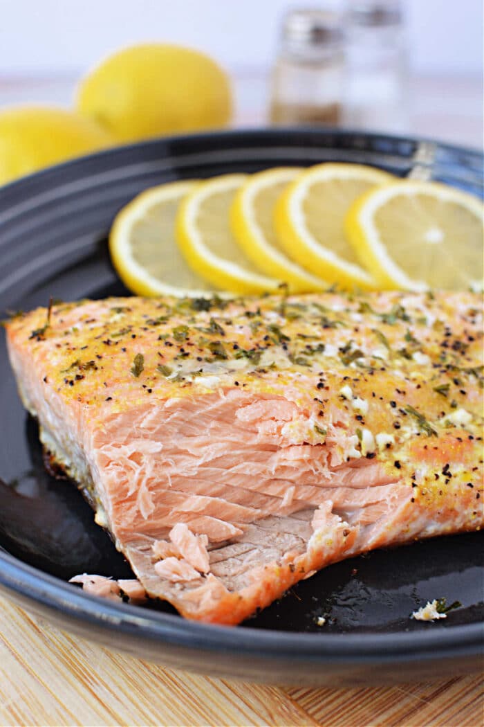 Frozen salmon baked in the oven, served on a black plate and garnished with herbs and lemon slices, with salt and pepper shakers behind.