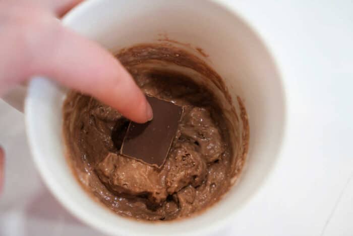 A hand presses a chocolate square into chocolate batter inside a white mug, preparing a quick and delicious Lava Cake.