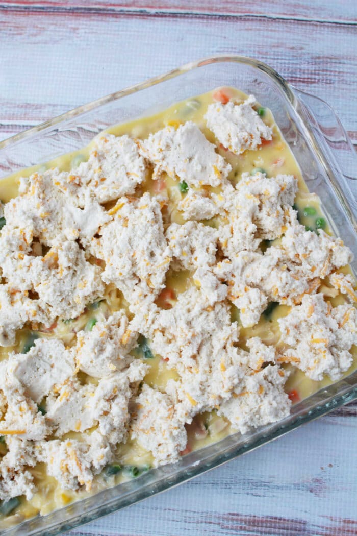 Unbaked chicken pot pie casserole topped with biscuits sits in a glass dish on a white wooden surface.