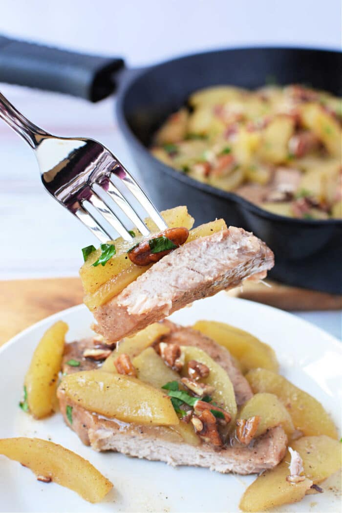 A fork holds a bite of baked pork chop topped with cooked apples and pecans above a plated serving and a skillet, perfect for showcasing this comforting Baked Pork Chops with Apples recipe.