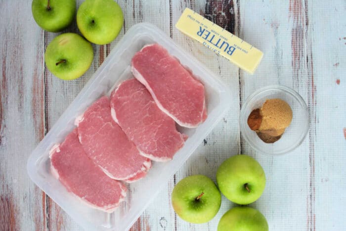 Raw pork chops in a plastic tray, green apples, a stick of butter, and brown sugar with spices on a table—perfect for trying a delicious baked pork chops recipe.