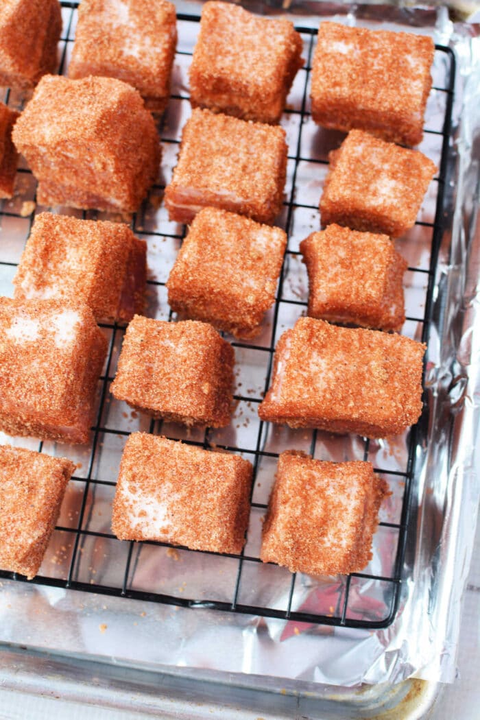 Squares of pork belly coated in a reddish-brown seasoning rest on a wire rack over a foil-lined tray, perfect for a savory burnt ends recipe.