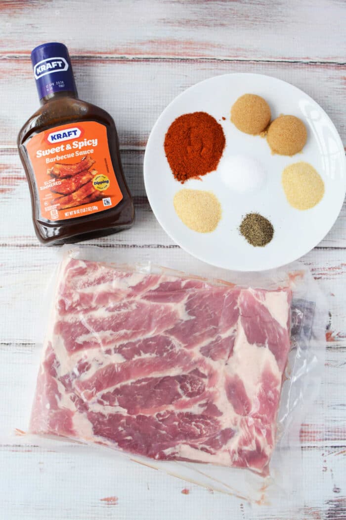 A slab of raw pork ribs, barbecue sauce, and a plate of six different spices await your next recipe on a white wooden surface&mdash;perfect for crafting smoky burnt ends or experimenting with pork belly delights.