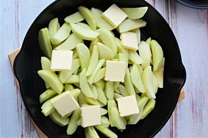 Sliced apples topped with butter pats in a black skillet on a light wooden surface, perfect for preparing Pork Chops with Apples or your favorite Baked Pork Chops Recipe.