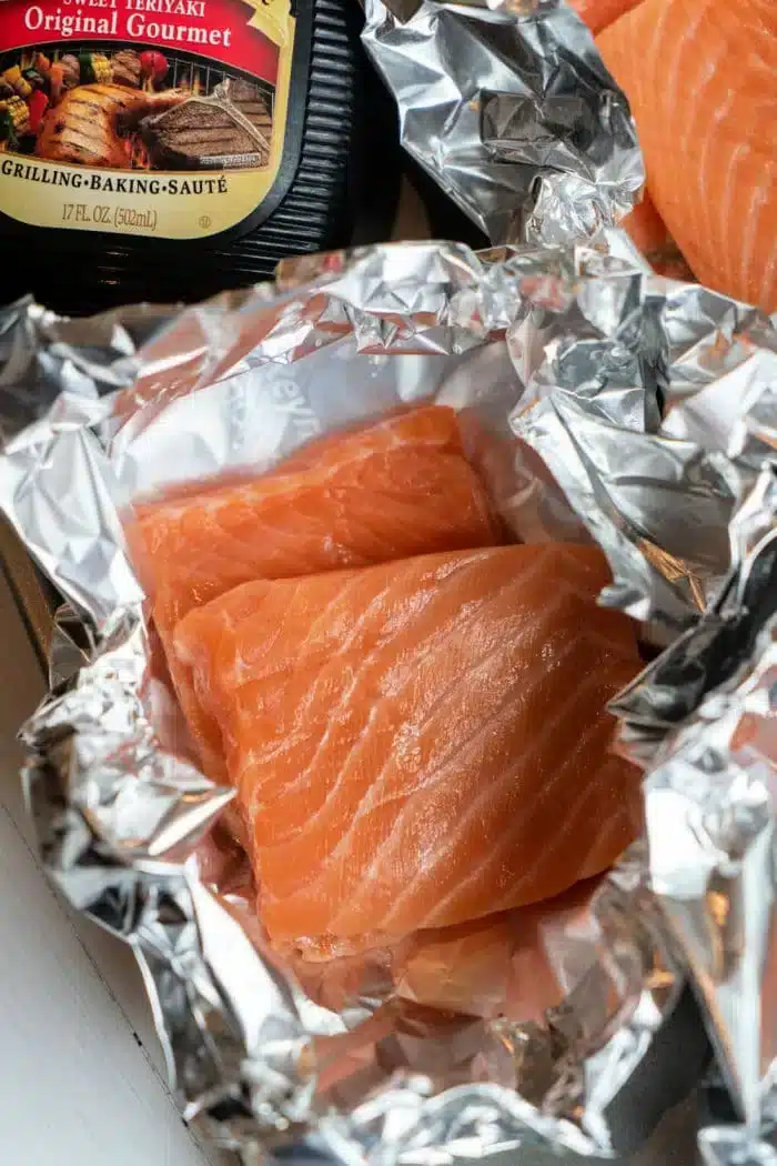 Oven Baked Teriyaki Salmon in Foil: Raw salmon fillets rest on aluminum foil with a bottle of teriyaki sauce in the background, ready for a flavorful oven-baked dish.