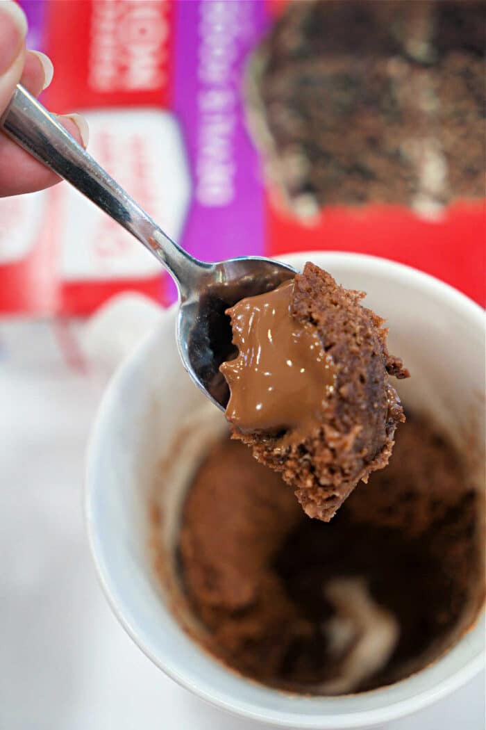 A spoon holds chocolate cake with a molten center above a white cup and cake mix box—perfect for your next Lava Cake Recipe or quick Mug Cake craving.