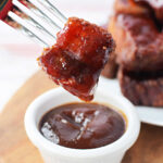 A fork holds a glazed pork belly bite above a small white bowl of barbecue sauce on a wooden board, perfect for any burnt ends recipe.
