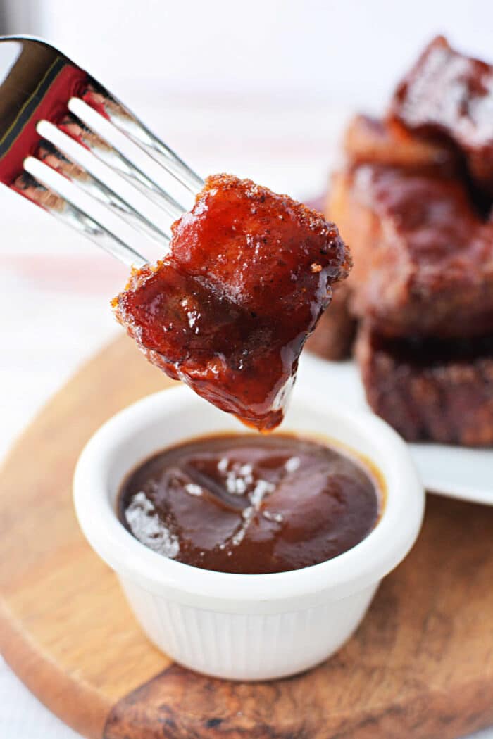 A fork holds a glazed pork belly bite above a small white bowl of barbecue sauce on a wooden board, perfect for any burnt ends recipe.