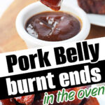 A fork holds a saucy pork belly burnt end over a bowl of BBQ sauce; more burnt ends, made from our delicious pork belly burnt ends recipe, are stacked on a plate.