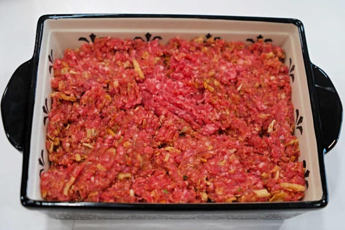 Raw ground beef mixed with onions is spread evenly in a rectangular baking dish, perfect for a classic meatloaf recipe and ready for cooking.