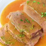 Sliced slow cooker beef bottom round roast with savory gravy and chopped parsley, served on a white plate.