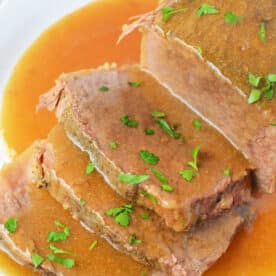 Sliced slow cooker beef bottom round roast with savory gravy and chopped parsley, served on a white plate.