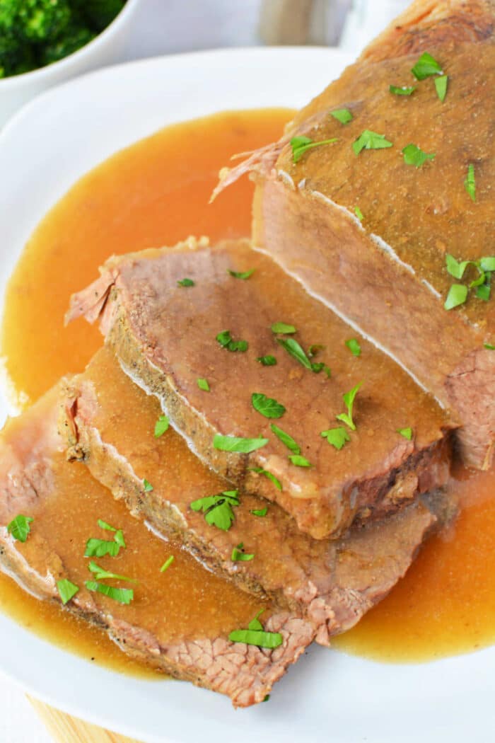 Sliced slow cooker beef bottom round roast with savory gravy and chopped parsley, served on a white plate.