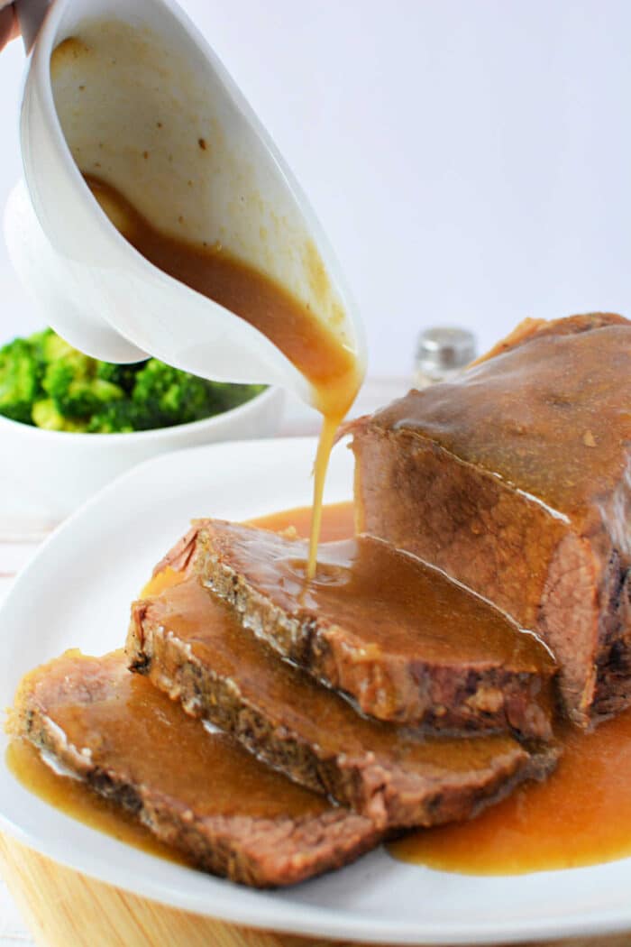 Slow cooker gravy being poured over sliced Beef Bottom Round Roast on a white plate, with broccoli in the background.