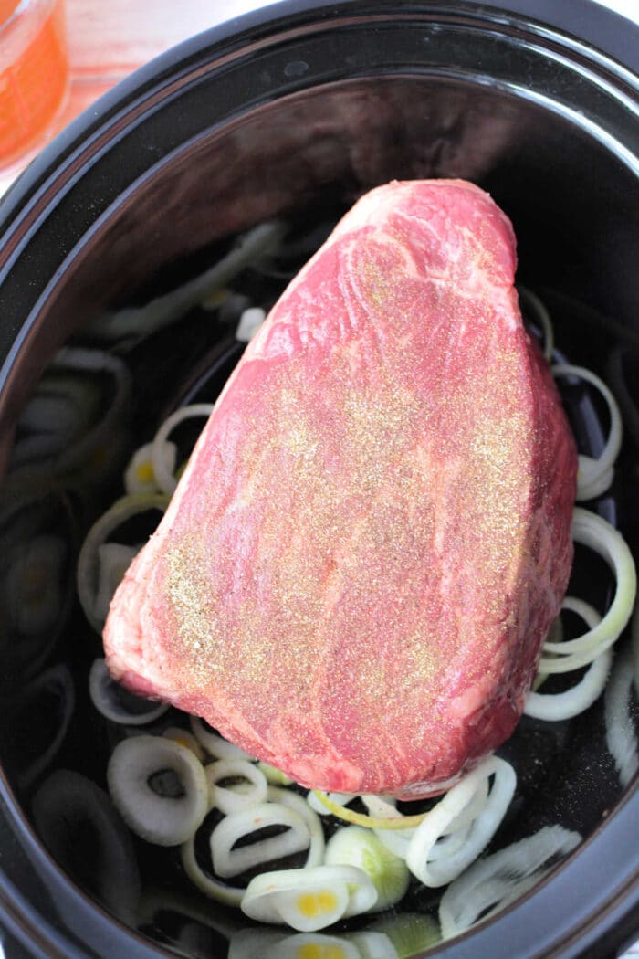 Beef Bottom Round Roast seasoned with spices sits on sliced onions in a black slow cooker, ready to be cooked.