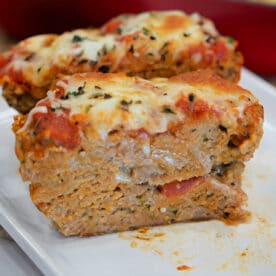 Two slices of cheesy, baked Chicken Parmesan Meatloaf topped with tomato sauce and herbs sit on a white plate.