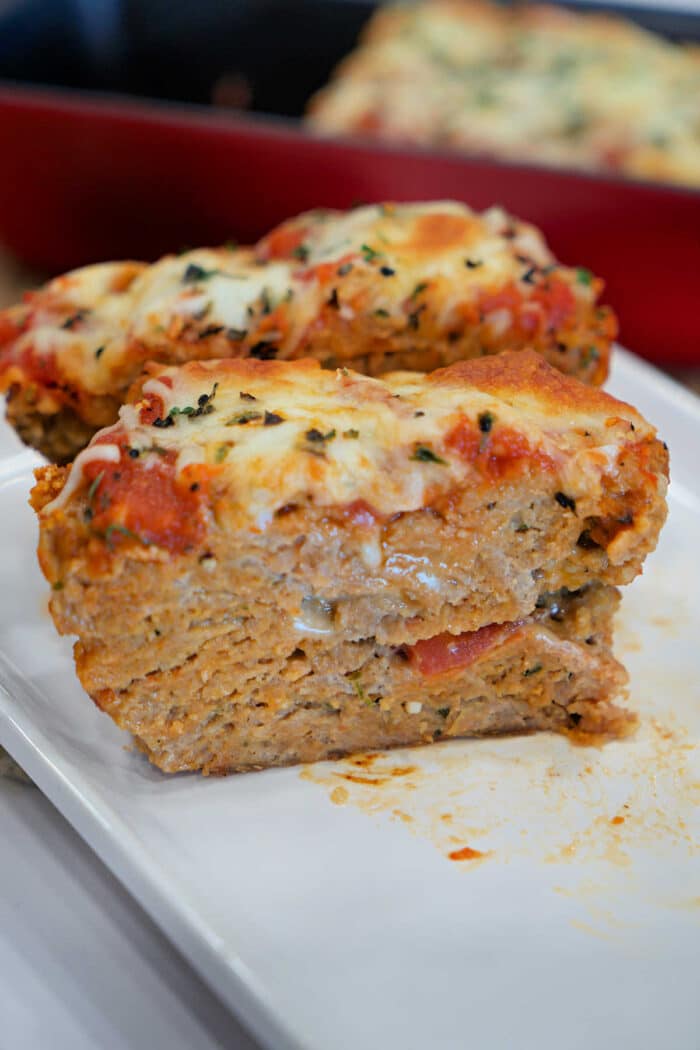 Two slices of cheesy, baked Chicken Parmesan Meatloaf topped with tomato sauce and herbs sit on a white plate.