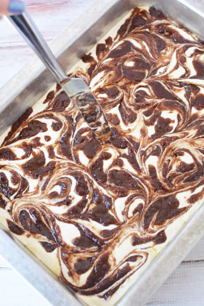 A hand swirls chocolate into batter with a knife in a metal baking pan, creating a marbled effect perfect for a Cinnamon Roll Cake Recipe.