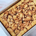A Cinnamon Roll Cake with a beautiful cinnamon swirl pattern, baked in a rectangular metal pan using an easy cake mix, rests on a light wooden surface.