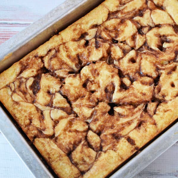 A Cinnamon Roll Cake with a beautiful cinnamon swirl pattern, baked in a rectangular metal pan using an easy cake mix, rests on a light wooden surface.