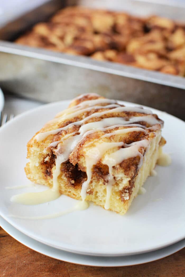 A slice of cinnamon roll cake made from cake mix with icing drizzle on a white plate, with more cake in a pan in the background.