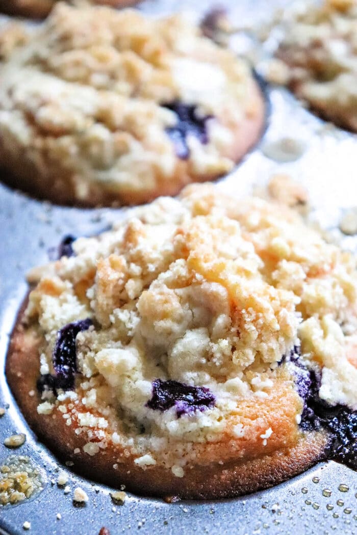 A close-up of a blueberry muffin with crumbly streusel topping in a metal muffin pan, showing how Blueberry muffin mix can improve texture for even better muffins.