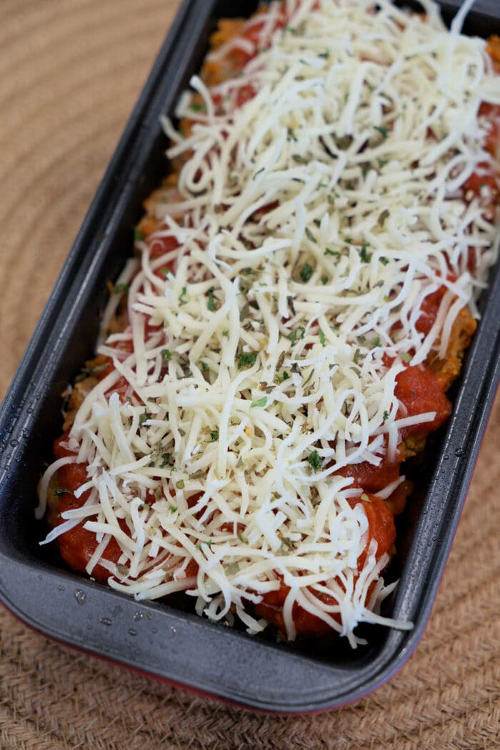 A loaf pan filled with Chicken Parmesan Meatloaf, topped with shredded cheese and herbs, ready to be baked.