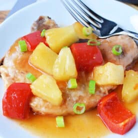 Tender pork chops and pineapple are topped with pineapple chunks, red bell peppers, sauce, and green onions, creating a vibrant dish served on a white plate.