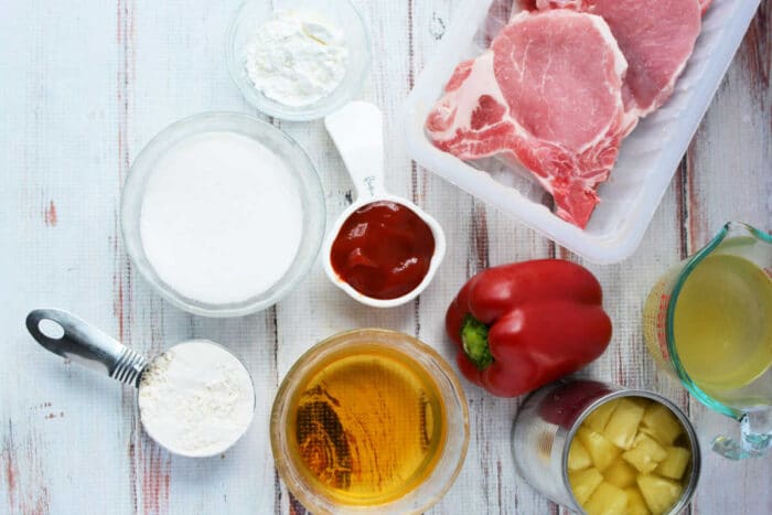Raw pork chops, red bell pepper, flour, sugar, ketchup, pineapple, broth, and cornstarch sit on a white surface—ideal ingredients for delicious Pork Chop Recipes.