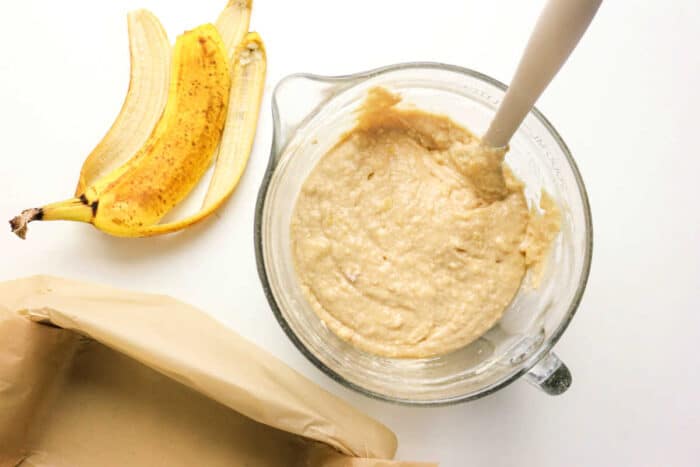 A glass bowl of Protein Banana Bread batter with a spatula sits beside banana peels and a parchment-lined pan, ready to be baked.