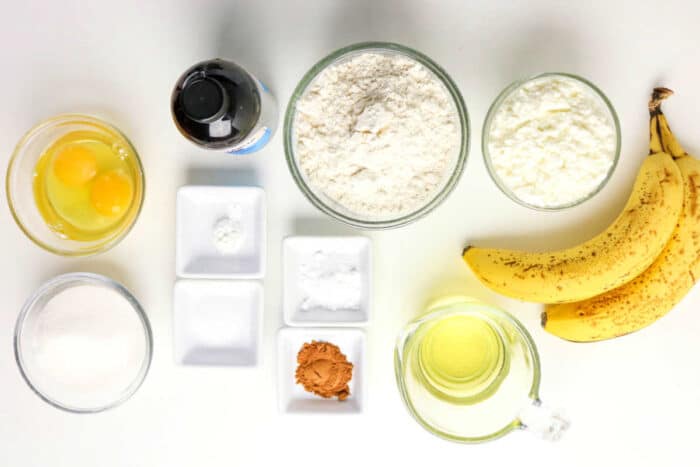 Top view of baking ingredients for Protein Banana Bread: eggs, flour, sugar, oil, bananas, vanilla, cinnamon, and small bowls with powders.