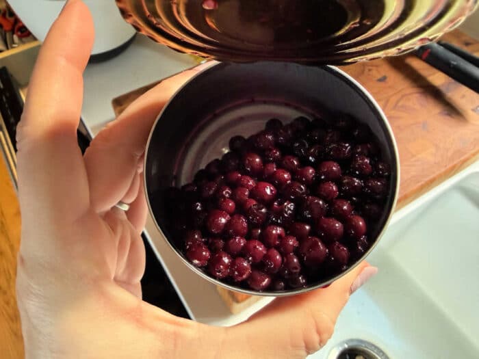 A hand holds an open can filled with dark purple berries over a kitchen sink and cutting board&mdash;perfect for learning how to make blueberry muffin mix better.