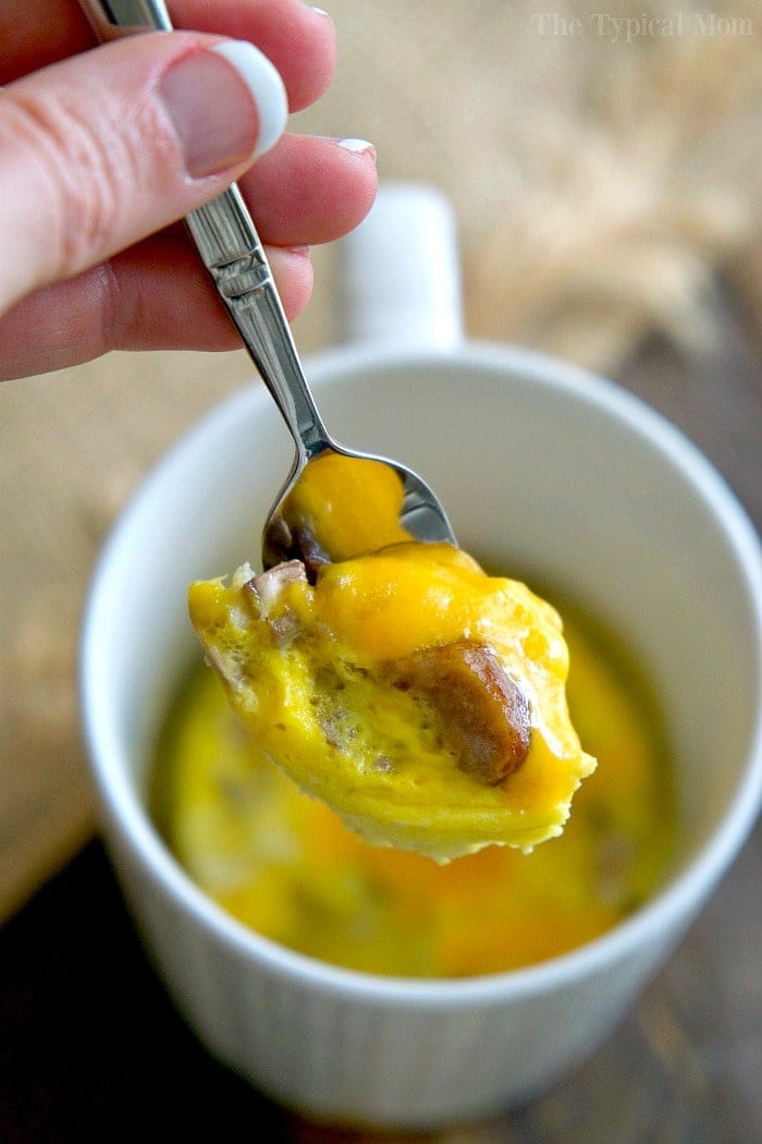 A hand holds a spoonful of cheesy egg and sausage from a white mug of microwave mug breakfast&mdash;perfect for fans of easy mug recipes.