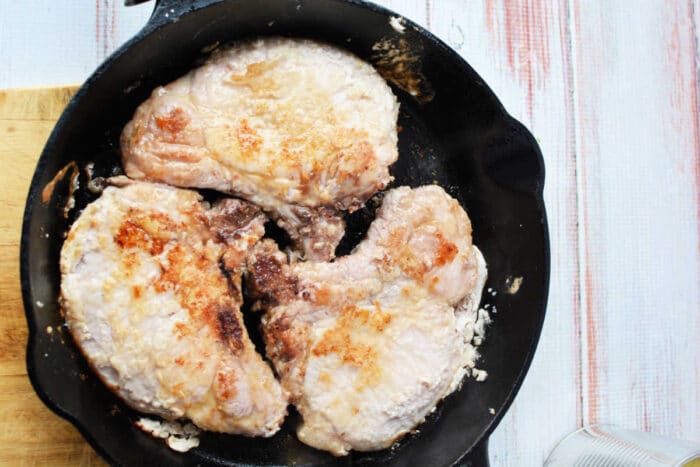 Four pork chops cooking in a black cast iron skillet on a light wooden surface, perfect for a savory pork and pineapple meal.