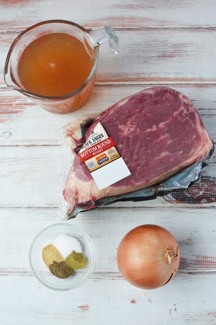 A raw beef bottom round roast, onion, spices, and beef broth in containers are arranged on a white wooden surface—perfect for your next slow cooker meal.