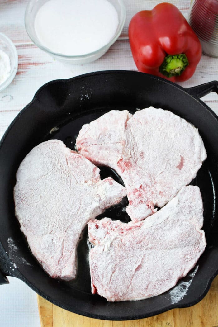 Three flour-coated raw pork chops in a black skillet, with a red bell pepper and a bowl nearby—perfect ingredients for trying new pork chop recipes.