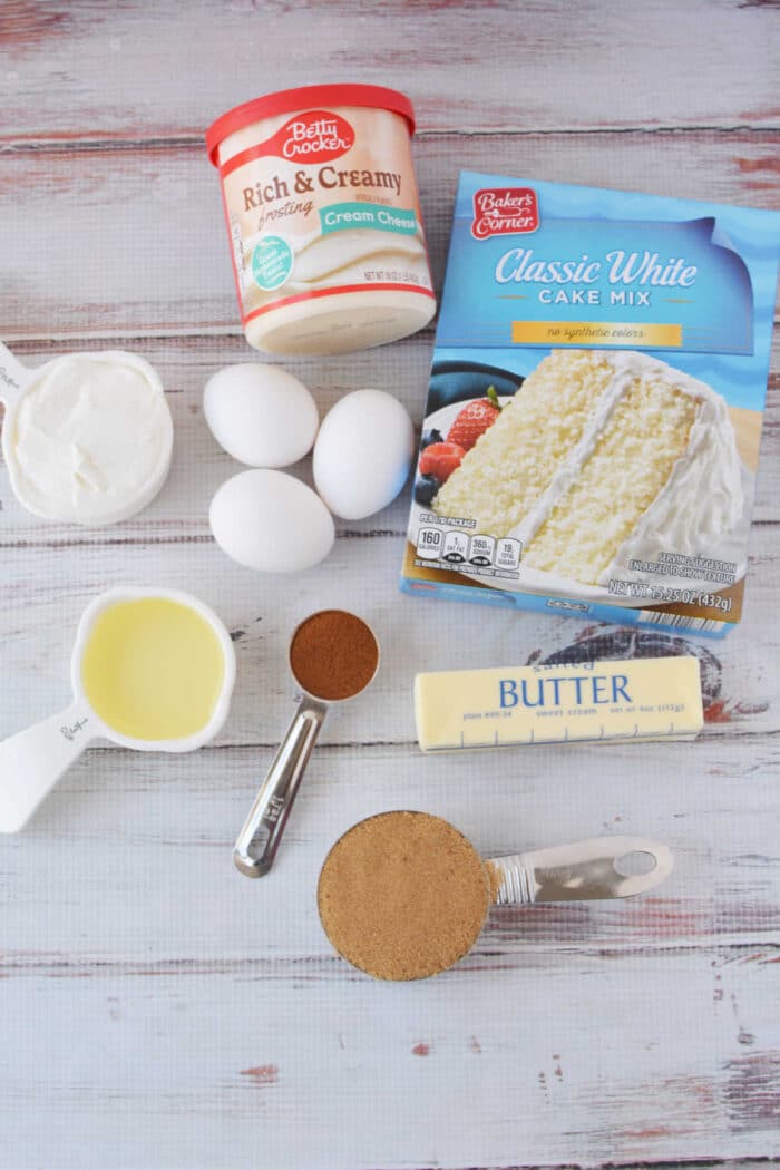 Cake mix, eggs, cream cheese frosting, butter, oil, sour cream, brown sugar, and cinnamon come together to create the perfect cinnamon roll cake on a white table.