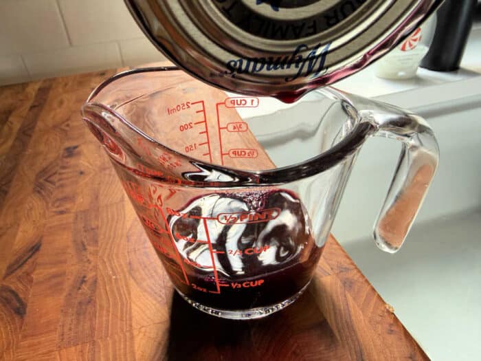 Red liquid being poured from a can into a glass measuring cup on a wooden countertop&mdash;perfect for learning how to make blueberry muffin mix better with flavorful additions.