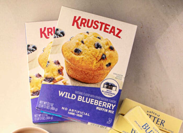 Box of Krusteaz Blueberry muffin mix on a counter next to butter packets and another muffin mix box, ready to help you improve your recipe.