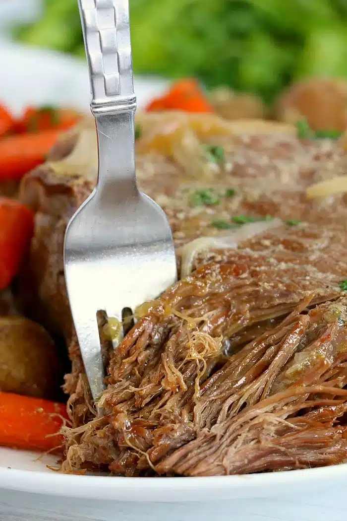 A fork shredding tender pot roast with carrots and potatoes on a plate, greens in the background&mdash;one of our favorite Ninja pressure cooker recipes.
