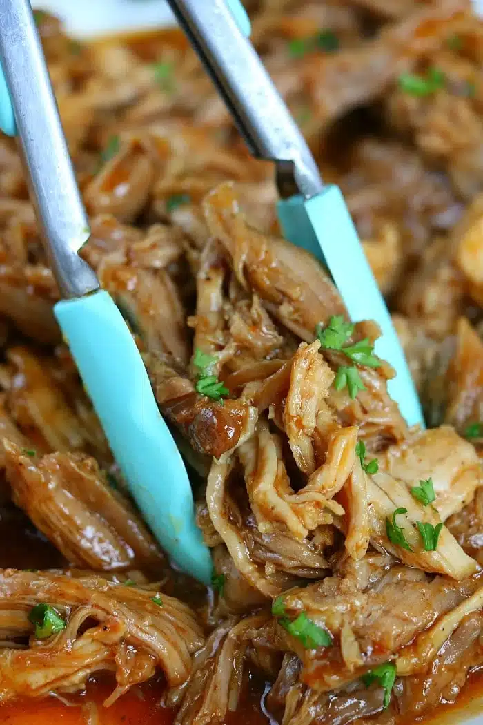 Tongs holding shredded, saucy pulled pork garnished with chopped parsley&mdash;perfect for trying out new ninja pressure cooker recipes.