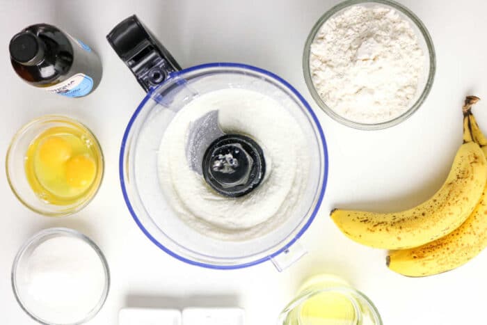 Top view of Protein Banana Bread ingredients: flour, eggs, bananas, sugar, oil, vanilla, and a food processor with flour inside.