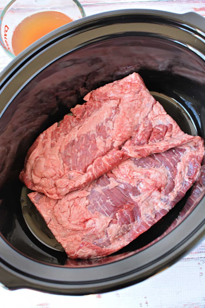Two raw pieces of beef, ideal for slow cooker skirt steak, are placed inside a black slow cooker, with broth in a measuring cup nearby&mdash;perfect for those seeking delicious skirt steak recipes.