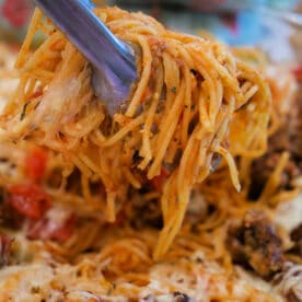 Tongs lifting cheesy baked spaghetti with meatballs from a casserole dish, showcasing gooey cheese and savory meatballs in every bite.