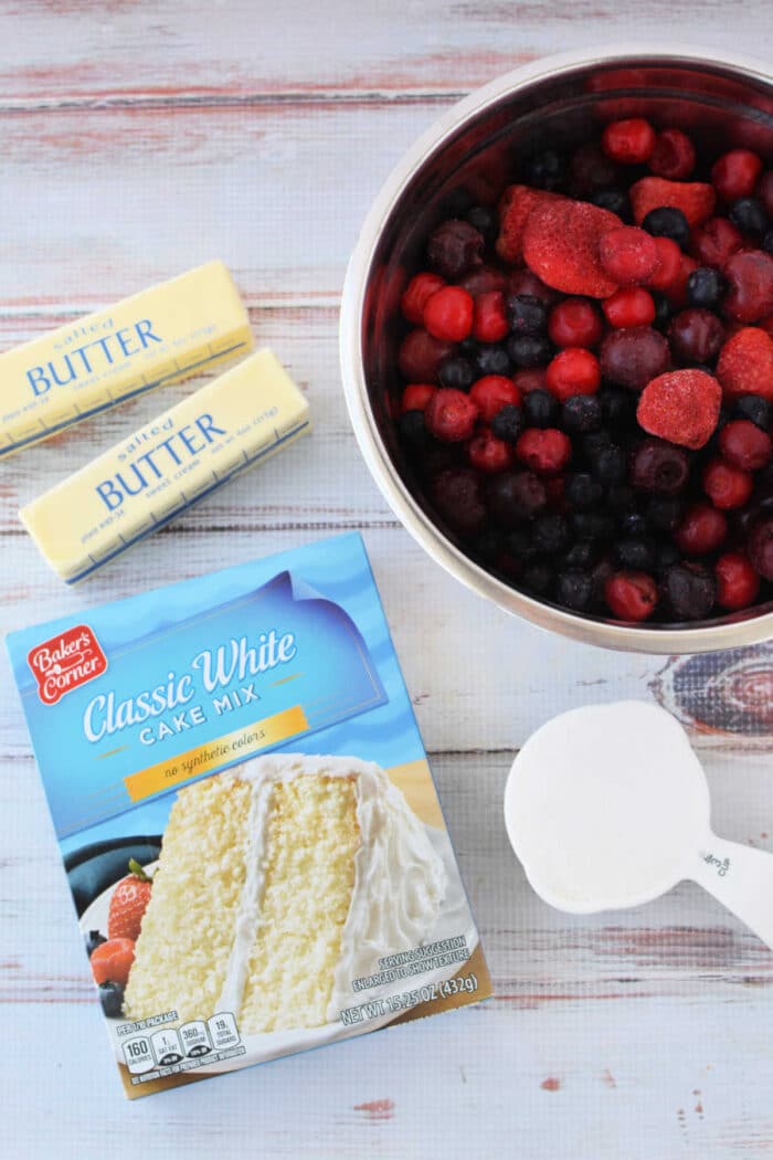 Box of cake mix, two butter sticks, frozen mixed berries in a bowl, and a cup of sugar on a white surface—perfect for making Berry Cobbler with Cake Mix.