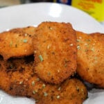 Golden, crispy breaded chicken cutlets on a plate, garnished with herbs, with Old Bay seasoning and a side of fried tilapia for the perfect tilapia recipe in the background.
