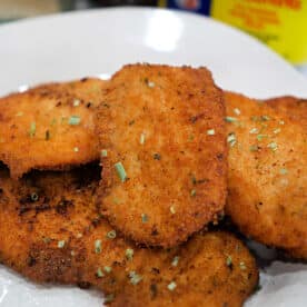 Golden, crispy breaded chicken cutlets on a plate, garnished with herbs, with Old Bay seasoning and a side of fried tilapia for the perfect tilapia recipe in the background.